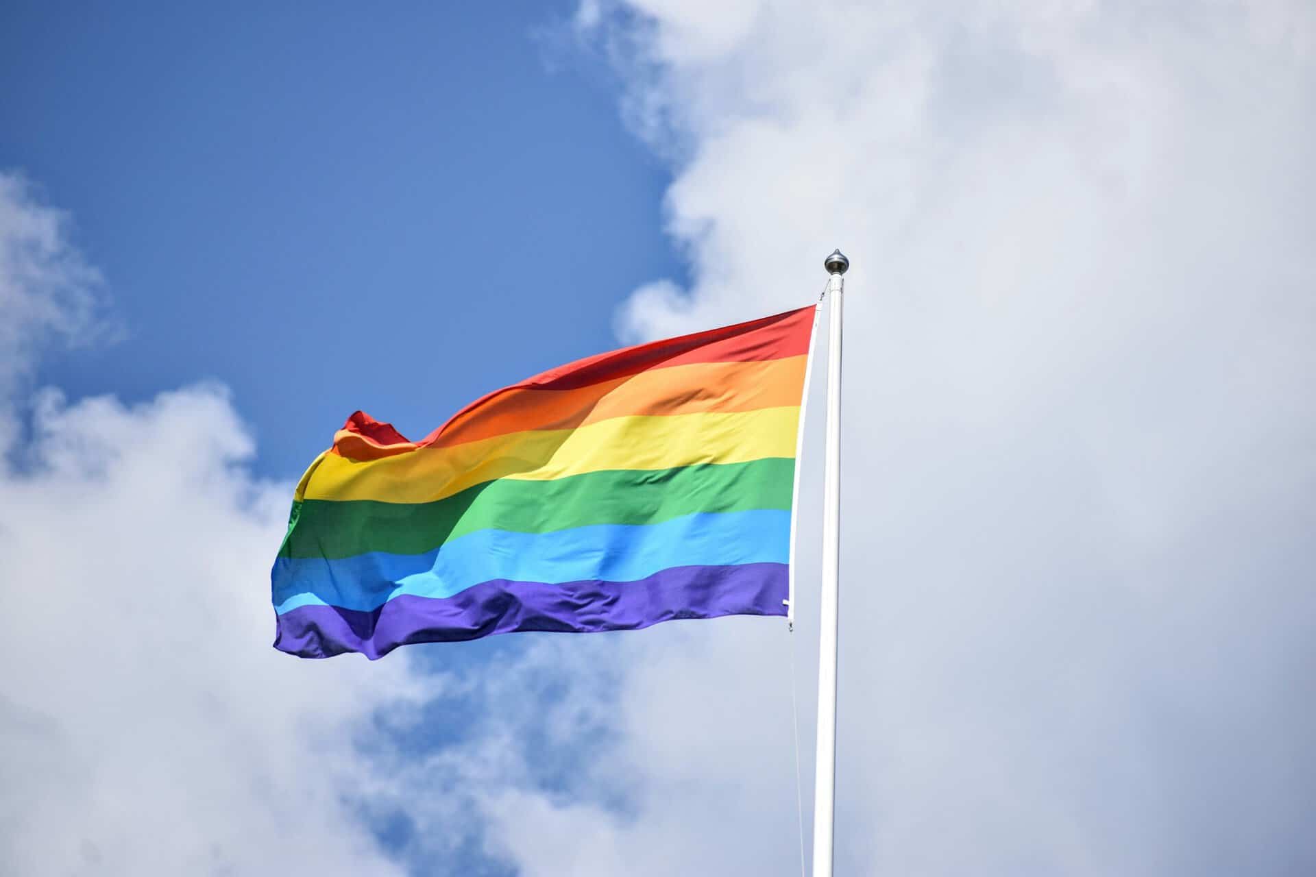 Rainbow Pride flag waving on a flagpole against a blue cloudy sky